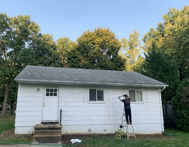 Technician pressure washing the exterior siding of a small white house from a ladder during exterior cleaning in Baytown